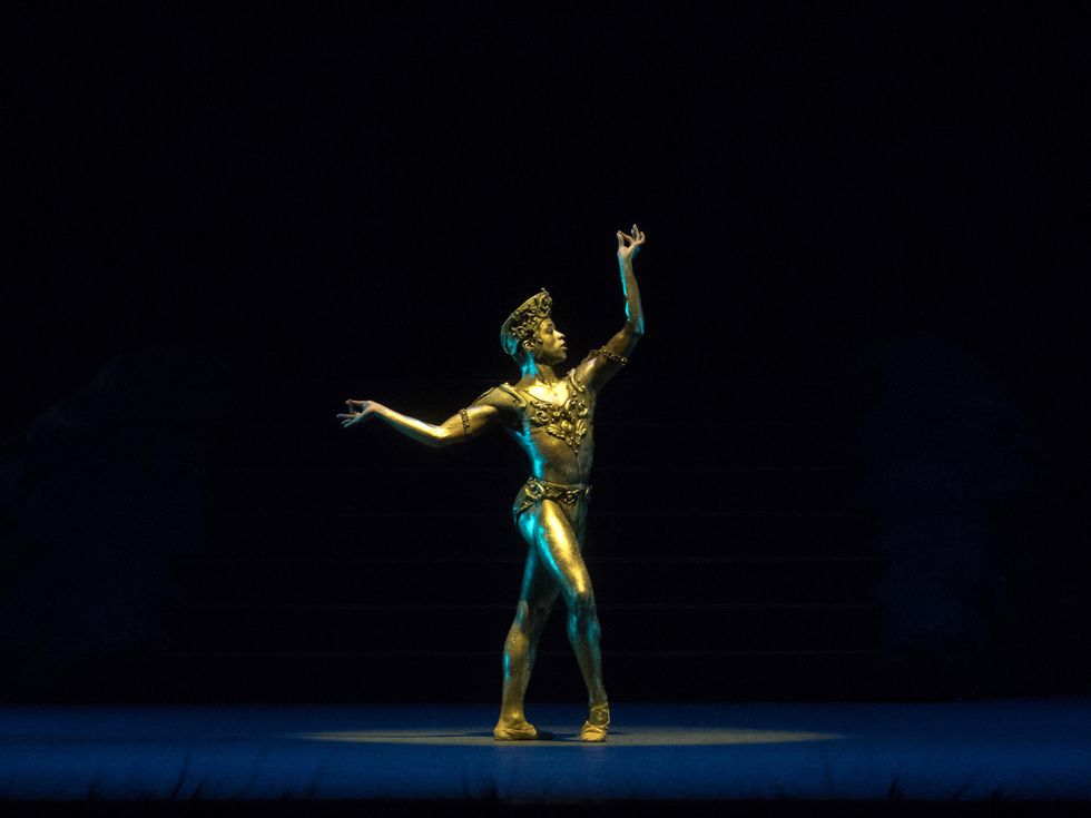 A male ballet dancer in gold body paint stands on a darkened stage bathed in a spotlight. He stands on his left foot and pops his right foot into demi-pointe, and bends his lifted arms at the elbows, pinching his thumb and forefingers together.