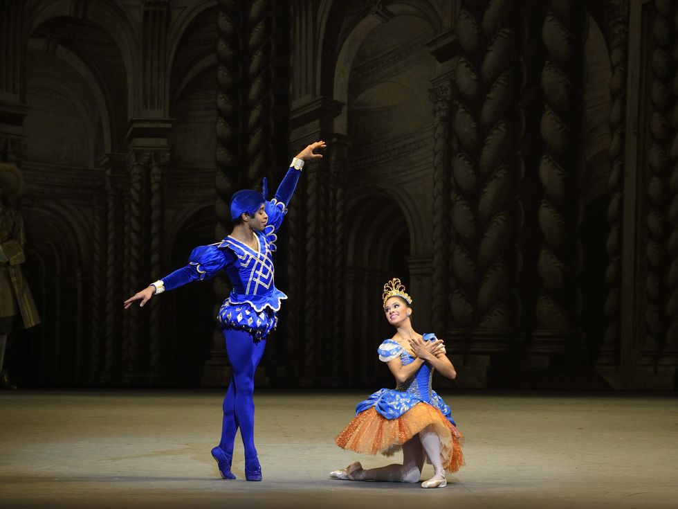 A male dancer with a blue feathered cap and wearing bright blue tights, shoes and tunic stands in sous-sus and lifts his arms like wings. To his left, a ballerina in a blue and orange tutu and gold crown kneels on the stage and crosses her hands at her chest. They look at each other and smile.