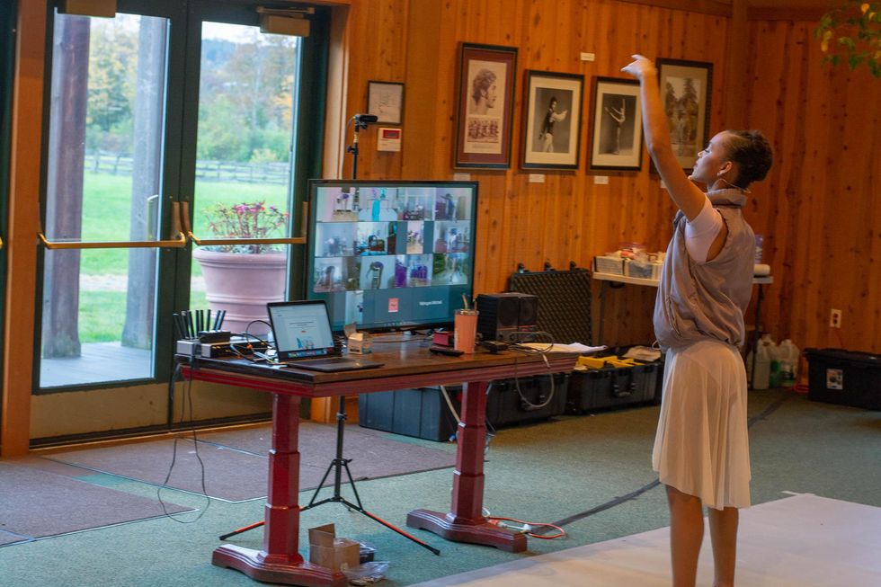 A female ballet teacher in a gray puffy vest, white leotard and skirt lifts her left arm up over her head and looks toward her hand in a spacious room with wood paneling. In front of her, a TV screen on a large table shows four rows of squares on Zoom.
