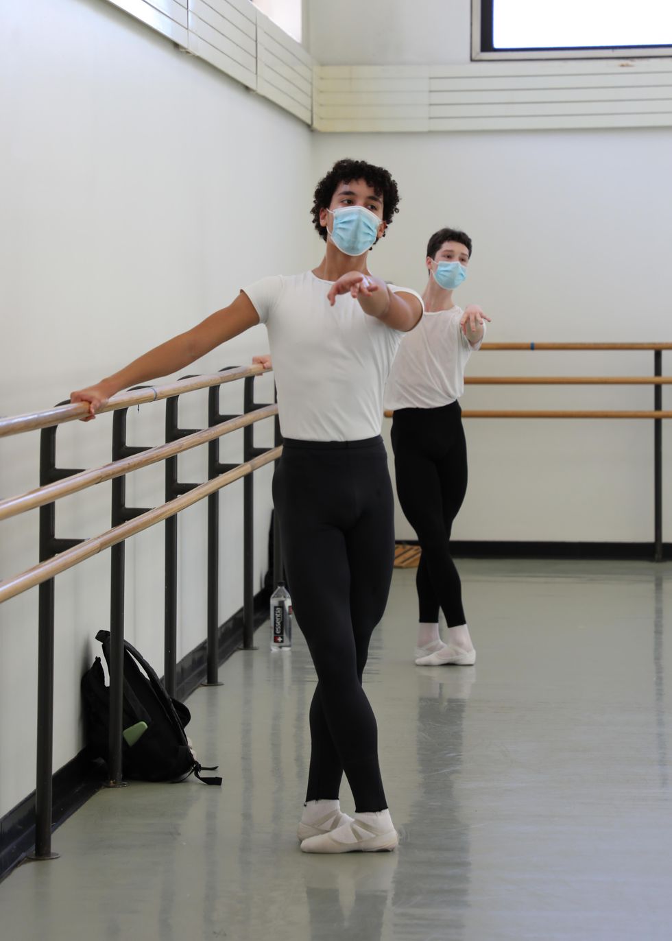 Two teenage boys in white T-shirts, black tights, white socks and slippers and blue facemasks stand at the barre in a tight fifth position. They hold onto the barre with their right hand and extend their left arms straight out in front of them.