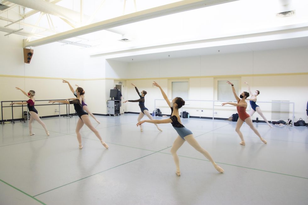 In a spacious, light-filled dance studio, two lines of teenage female ballet students do a tendu derriere effac\u00e9 in pli\u00e9 on their right leg with their arms in third arabesque. They each wear a leotard in various colors, pink tights and pointe shoes.