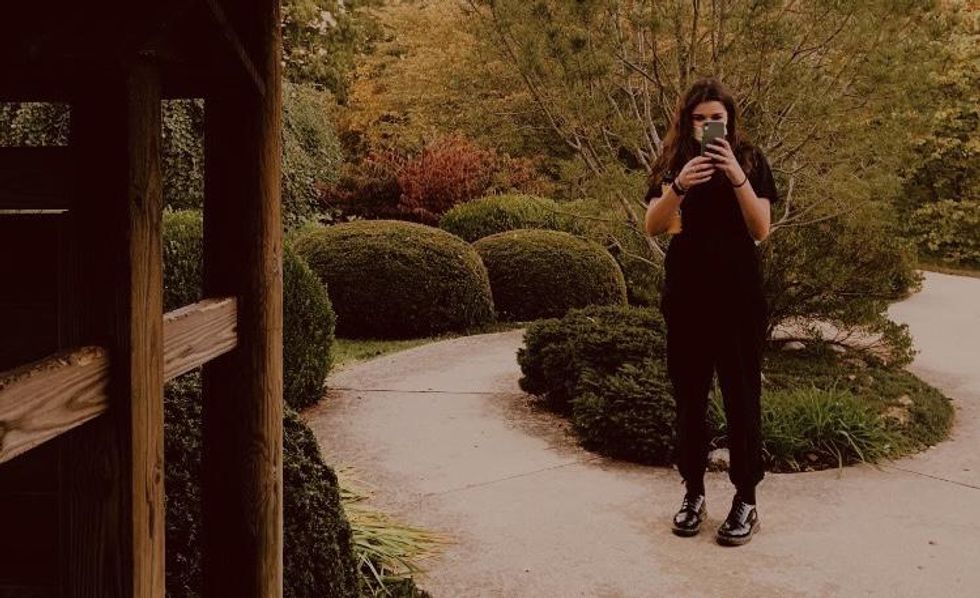 Fashionable girl standing in garden taking a picture of the person behind the camera.