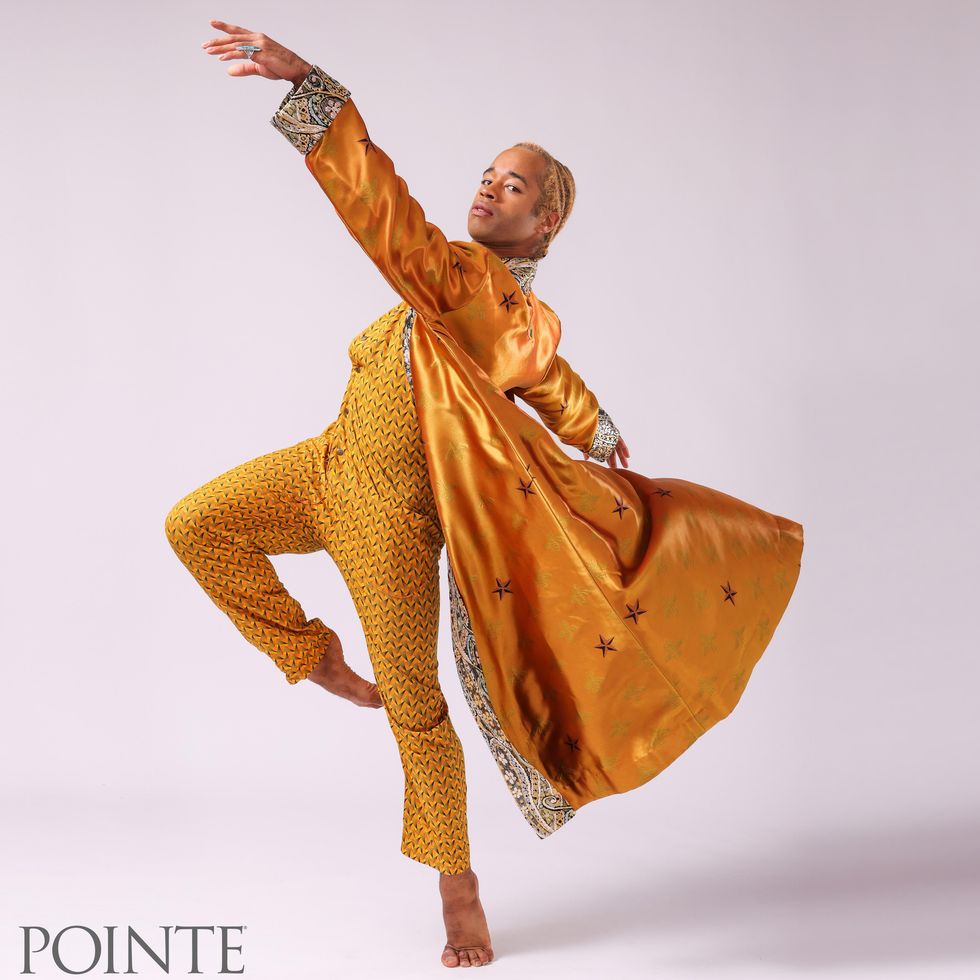 Gabe Stone Shayer wears a shiny, floor-length gold overcoat and yellow one-piece jumper with a black zig-zag pattern. He does a turned in pass\u00e9 relev\u00e9 on his left leg and crosses his left arm high across him, which makes his jacket float up behind him.