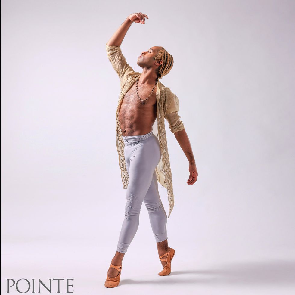 Wearing white tights and a long, sheer off-white coat, a Black male ballet dancer stands in fourth position on demi-pointe and looks up towards his lifted right hand.