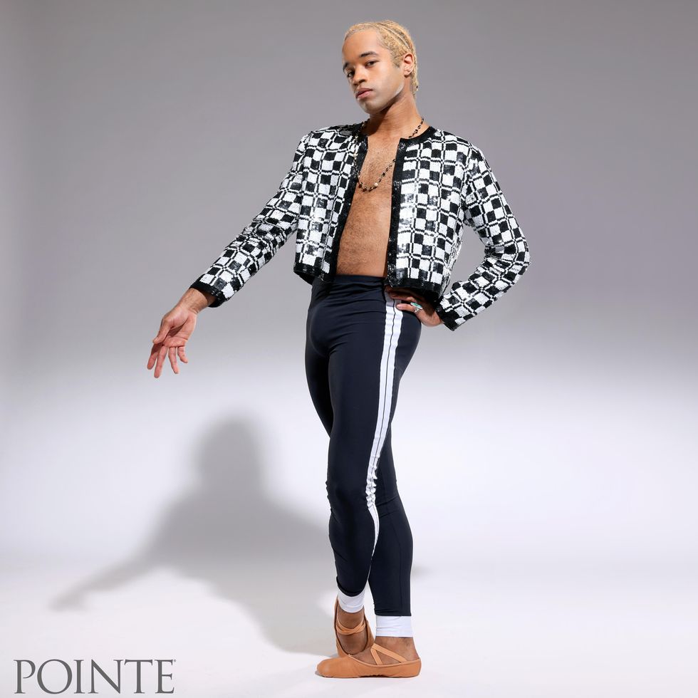 A male, light-skinned Black ballet dancer stands on his right foot and crosses his left foot over it while holding his left hand on his hip and his right hand out pointing towards the ground. He wears a sparkly white and black bolero jacket over his bare chest, a beaded necklace and black leggings with a white stripe down the sides.