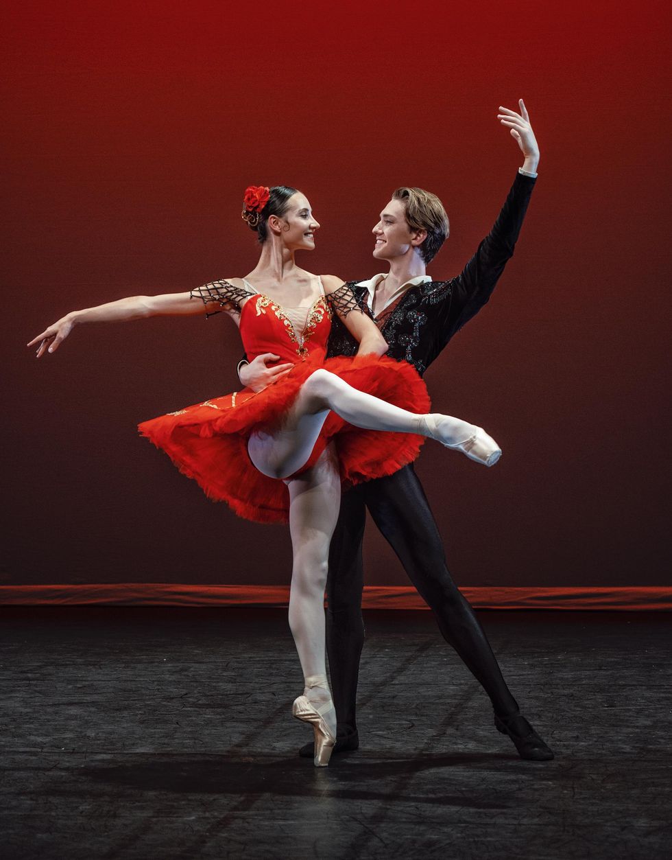 A white ballerina in a bright red tutu, pink tihgst and pointe shoes and a rose in her hair performs an attitude devant with her right leg and looks towards her partner on her left. 