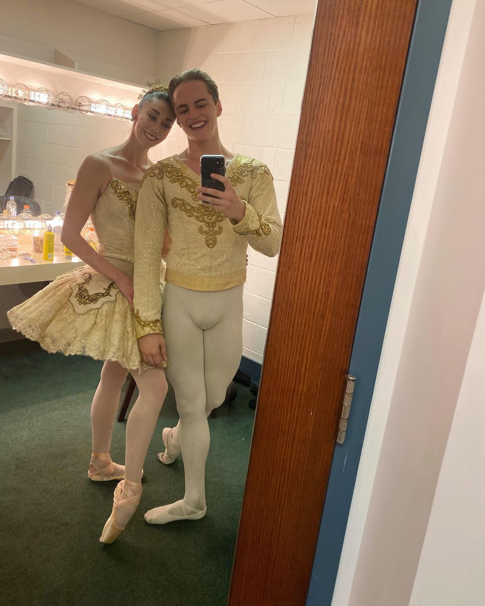 A male and female dancer stands next to each other closely and pose for a selfie in a green-carpeted dressing room. 