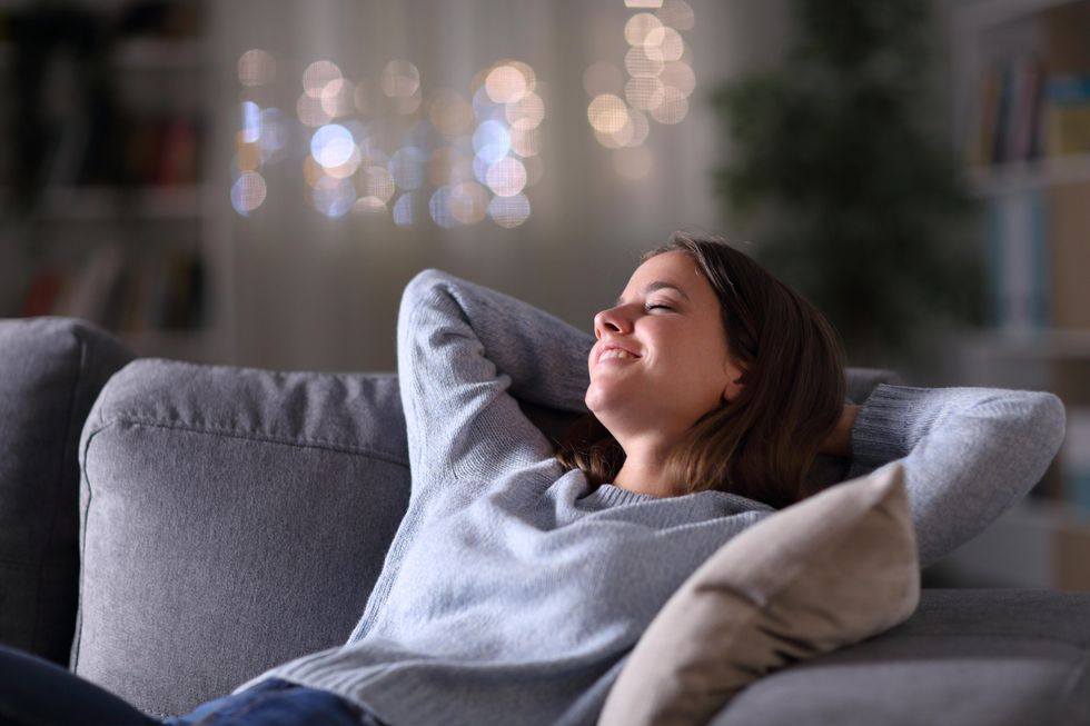 A white woman with straight brown hair reclines on a coach with her eyes closed. She is smiling. In the blurred-out background, there is a Christmas tree.