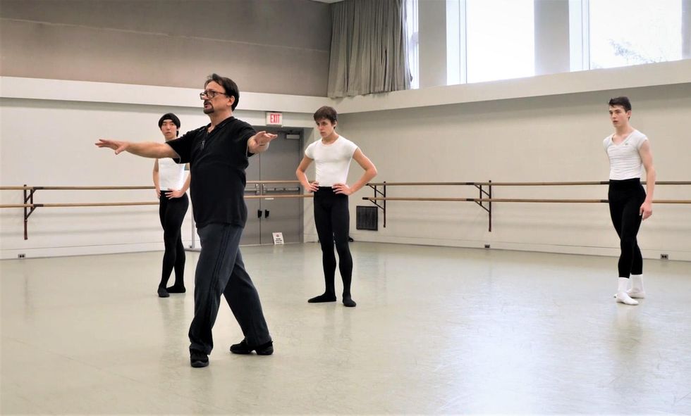Dhouloukhadze, a man with a goatee and black shirt and pants, demonstrates a fourth position prep while three teenage boys look on in a ballet studio.