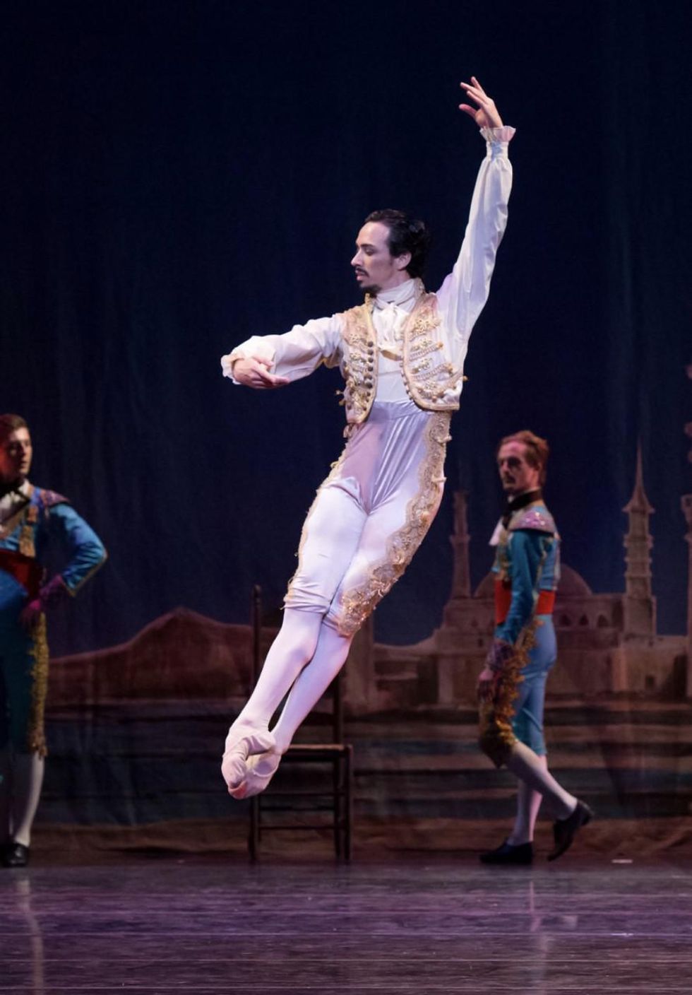 A white male danseur in a white and gold Spanish-style bolero jacket and tights, is shown onstage doing an assembl\u00e9 to the right.