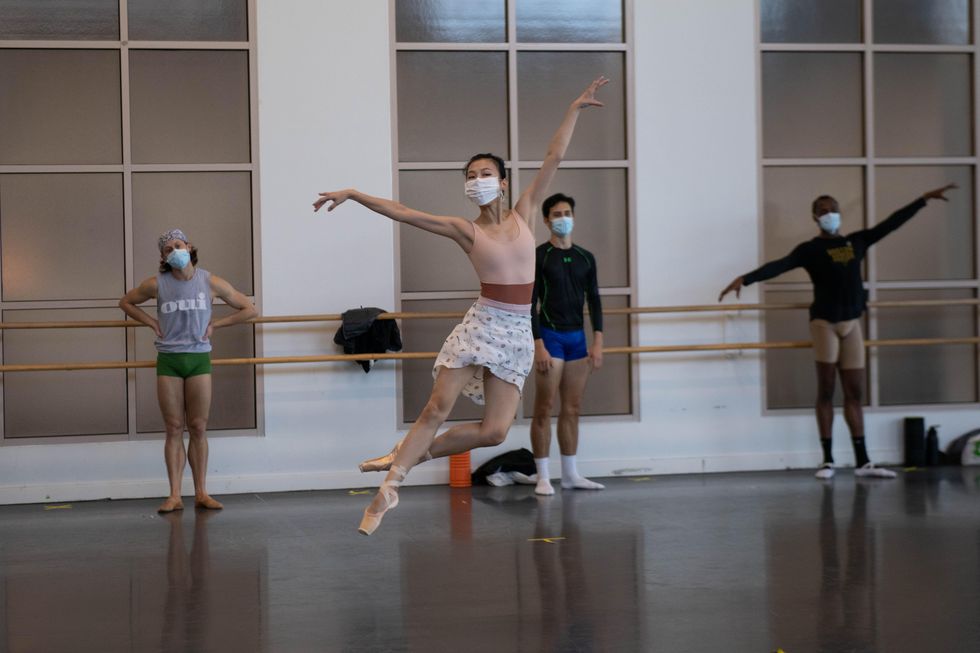 An Asian ballerina in a face mask, pink leotard, white skirt and pointe shoes does a jet\u00e9 saut\u00e9 with her left foot back in coup\u00e9. Behind her three male dancers in practice clothes wait near the barre.