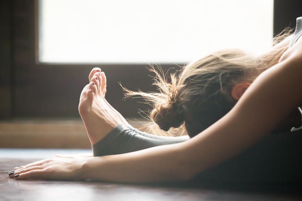 A young athletic woman with gray leggings and her hair in a messy bun bends over her outstretched legs on a yoga mat.
