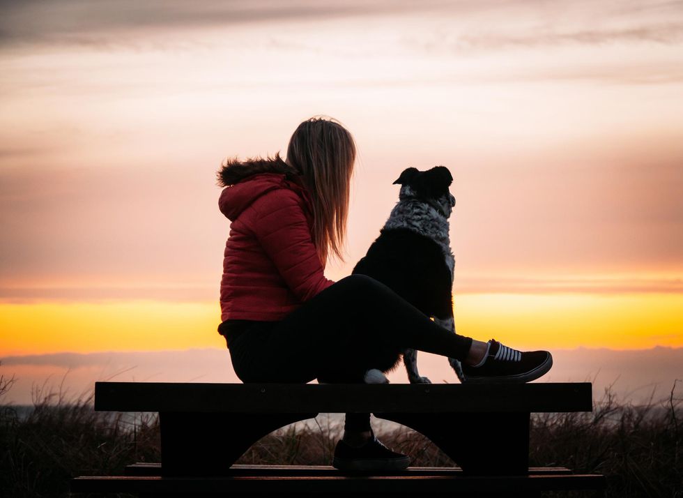 A long-haired woman wearing a red hooded puffy coat, leggings and sneakers sits on a beachside bench with her dog and watches the sunset.