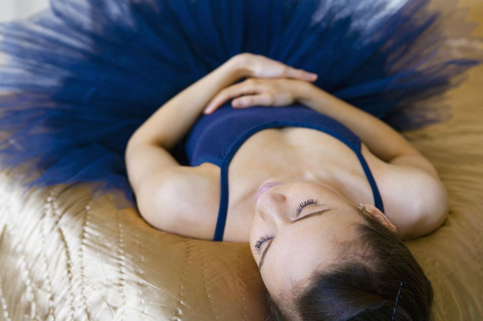 A teenage ballet student in a navy blue leotard and tutu rests on a bed with her eyes closed and hands on her stomach.