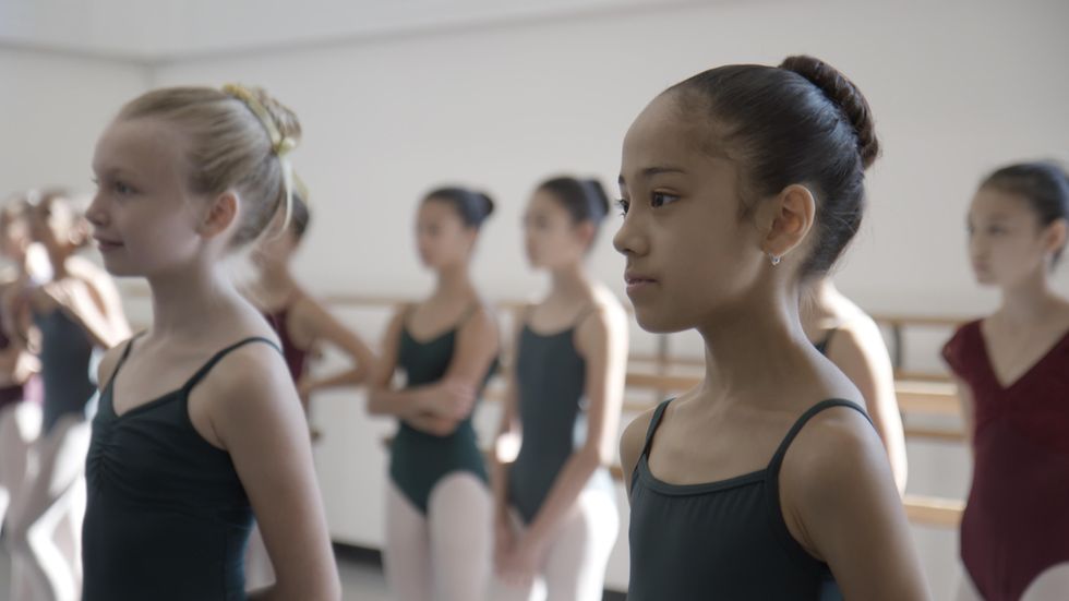 Two girl ballet students wearing black camisole leotards and their hair in a bun stand next to each other in a brightly lit dance studio, listening attentively as someone speaks to them off camera. Seven other young girls dancers in leotards and upswept hair stand casually behind them.