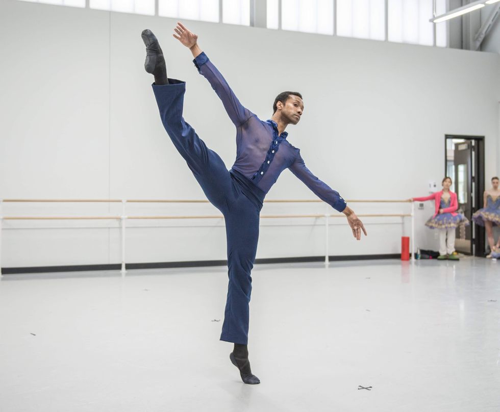 An African American male dancer, wearing indigo pants and a mesh, indigo button-down blouse, does a grand battement with his right leg and lean off balance to the left, with his arms out and tilted in a diagonal.