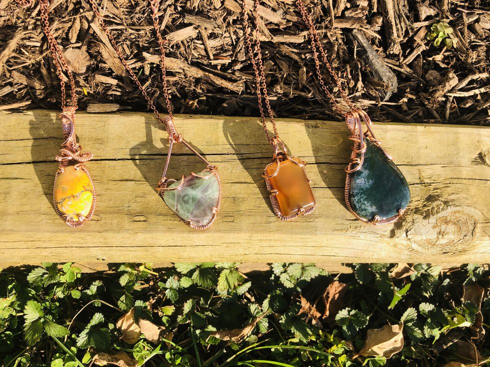 Four copper-wire necklaces with a yellow, light green, orange and dark green stone, respectively, are lined up right to left on a plank of wood.