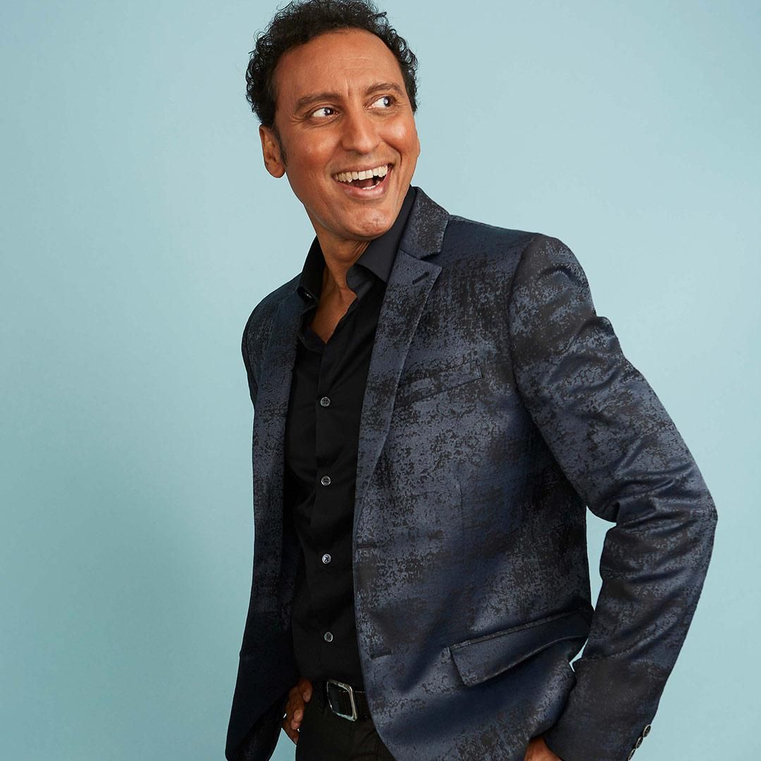 Evil star Aasif Mandvi smiles and looks over his shoulder in front of a soft blue wall