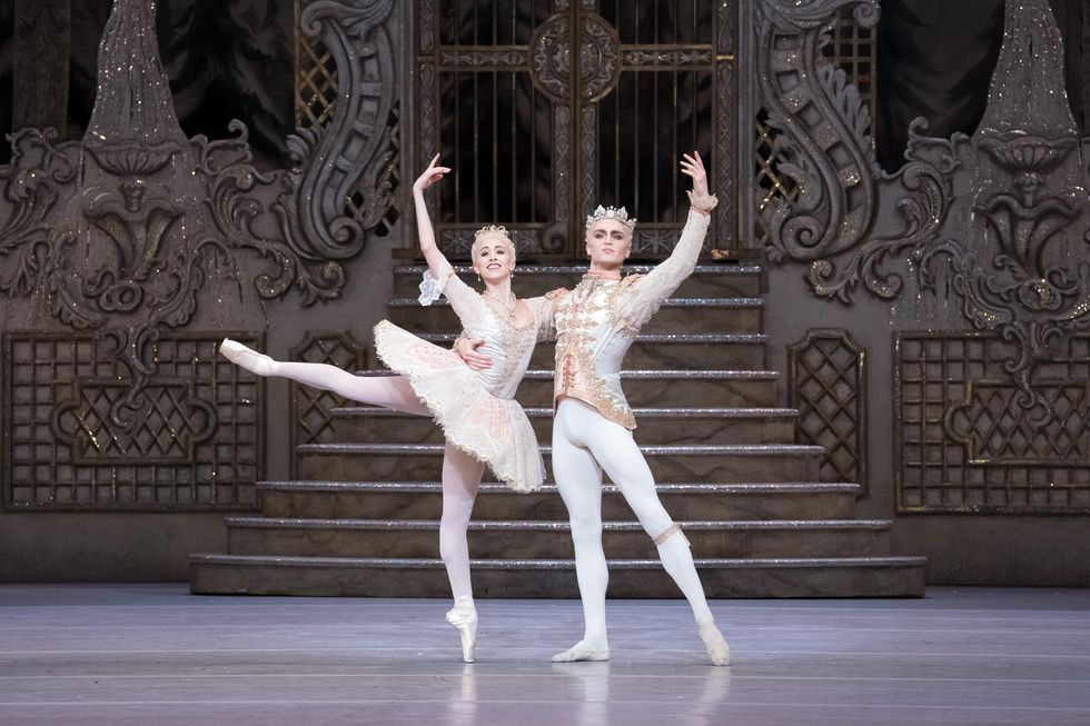 A ballerina in a tiara and light pink tutu, tights and pointe shoes stands in attitude with her right arm up and her left arm around her partner's right shoulder. He stands in tendu and wears a matching jacket and tights and crown.
