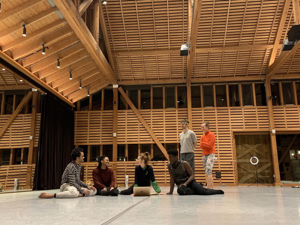 A group of of four dancers sit on the ground, with two other dancers standing behind them to the right. They are in a large dance studio with wood-planked walls, and the woman sitting in the middle of the group has a silver laptop.