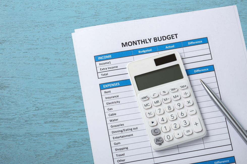 A small white calculator and silver pen sits on top of a pile of papers labeled "monthly budget." The budget sheets are divided into grids labeled "income" and "expenses," with categories for living expenses like rent.