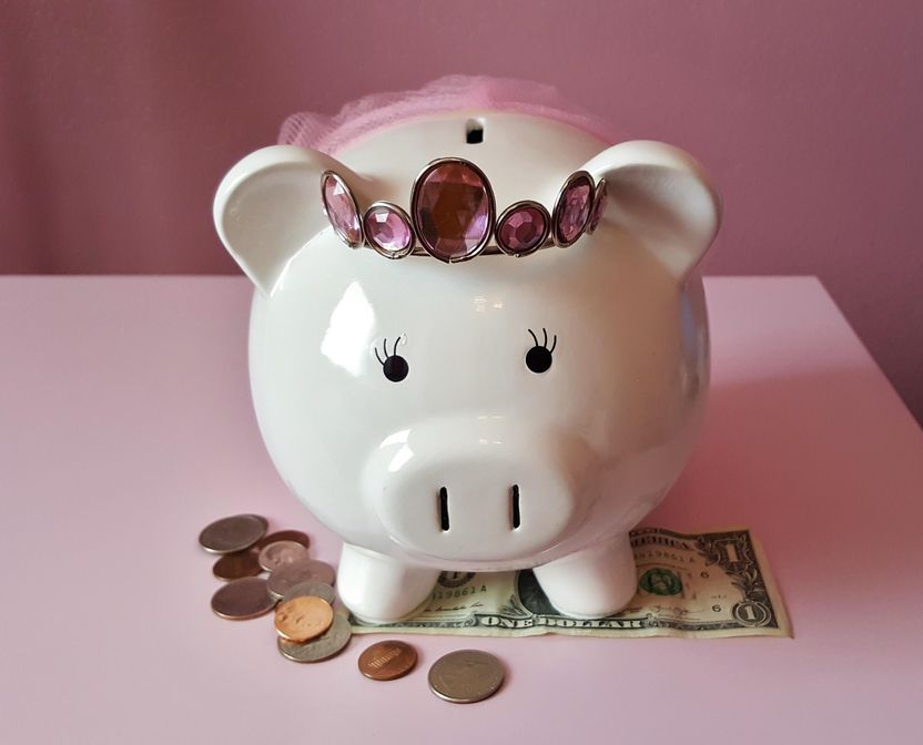 A cute white ceramic piggy bank wears a jeweled tiara and pink tutu, and stands on top of a dollar bill and next to a pile of coins.