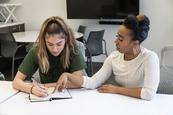 Chanel DaSilva sits at a white table with a teenage girl, pointing at a notebook the girl is writing in. There are other tables and chairs and a TV in the background.