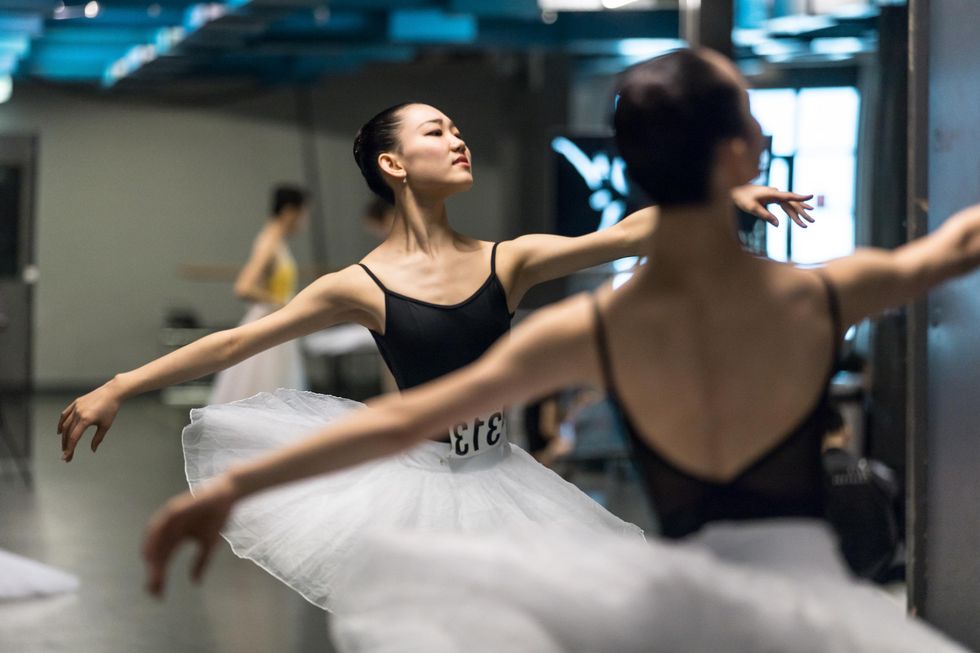 A female Asian ballet student in a black leotard and white tutu practices her port de bras in front of a mirror backstage.