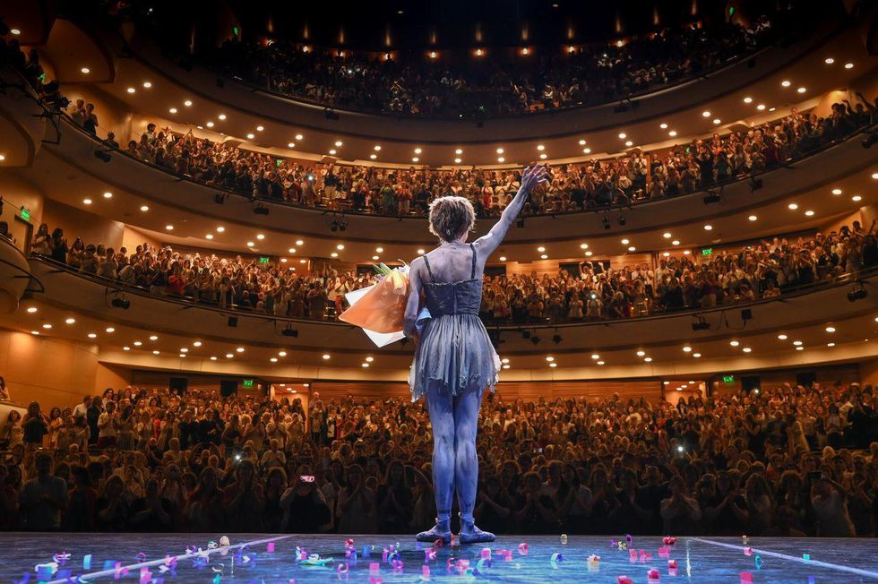 In this photo shot from behind, Mar\u00eda Riccetto wears the ragged dress costume and short wig from the ballet Manon and stands center stage, holding a large bouquet of flowers and waving to the audience. The packed crowd is on its feet, cheering her final bow.
