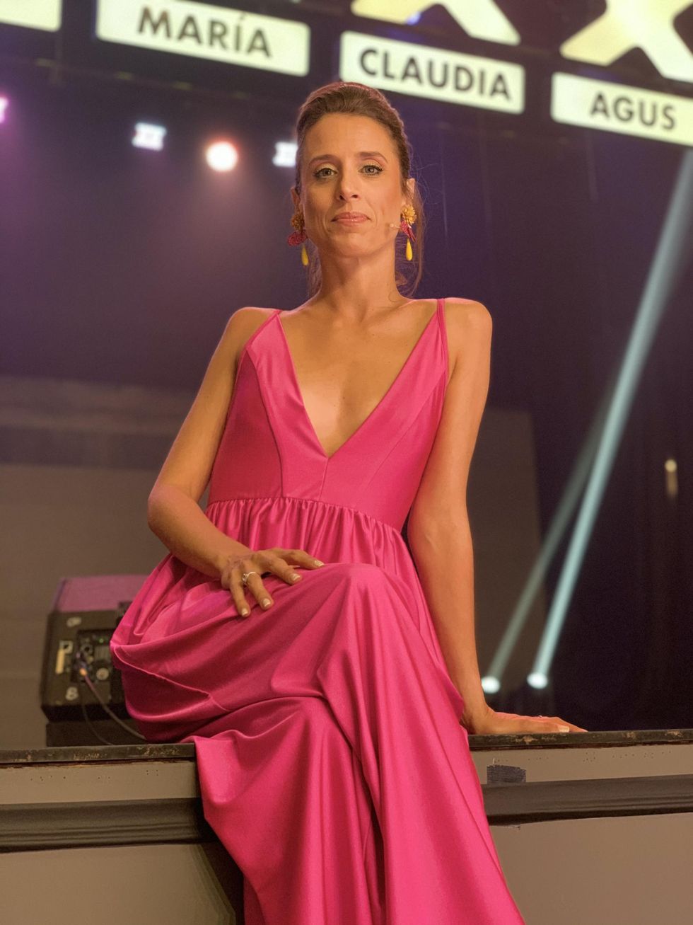 Wearing a bright pink gown with a plunging V-neckline, Mar\u00eda Riccetto sits cross-legged on the television stage of Got Talent Uruguay and smiles confidently towards the camera.