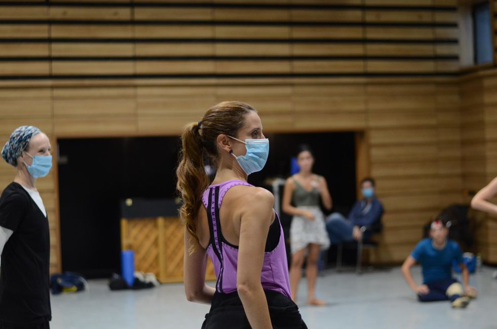 With her brown hair in a ponytail, Mar\u00eda Riccetto wears a blue face mask and looks to her right, directing a group of dancers who also wear masks in a large dance studio.