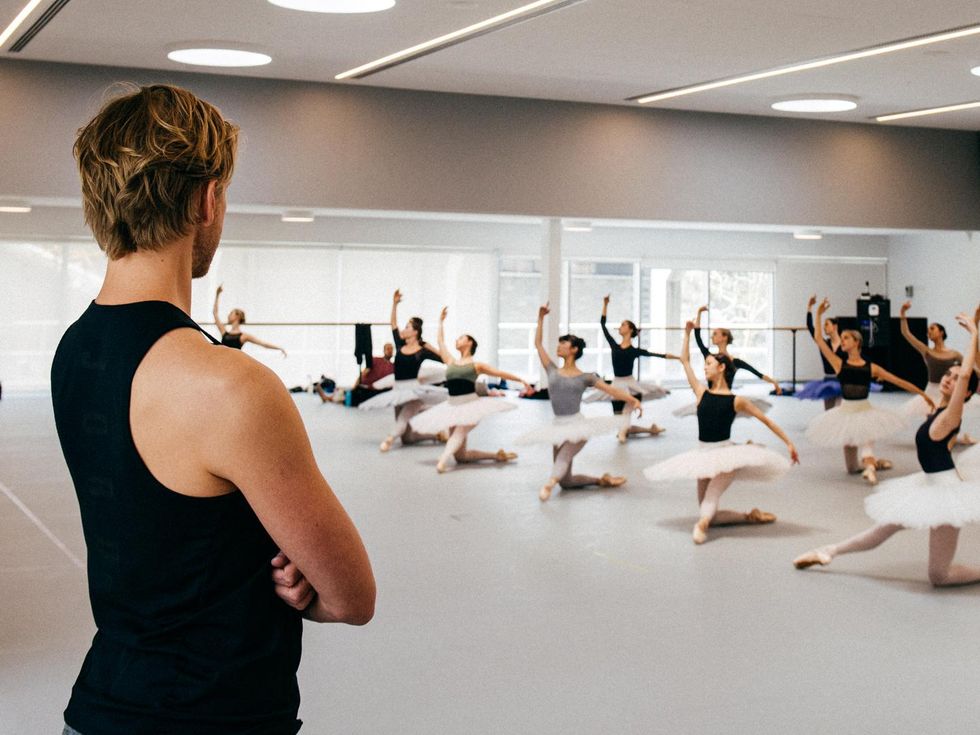 A blond David Hallberg is shown from the back wearing a black tank top, watching rehearsal with his arms crossed. In front of him in the dance studio, three lines of ballerinas kneel on their right knee with their left leg extended and cambr\u00e9 slightly back with their arms in third position. They each wear a leotard, pink tights, pointe shoes and a white practice tutu.