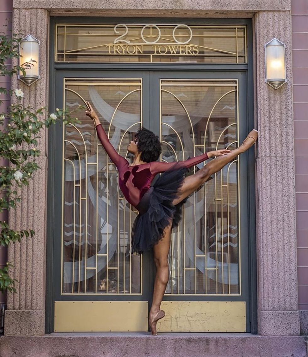Alexandra Hutchinson does an arabesque on pointe in front of a NYC building's art-deco style entrance. She is wearing a maroon, mesh and velvet leotard, a black tutu, bare legs and flesh-colored pointe shoes.