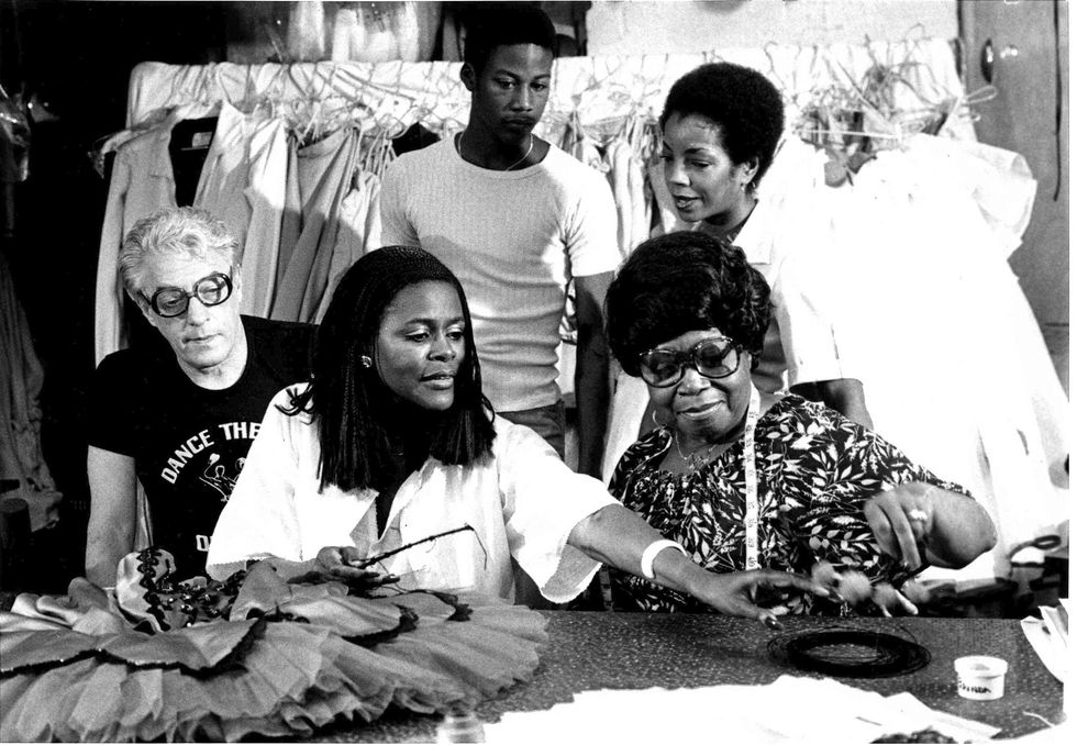 Cicely Tyson, wearing a billowy white blouse and her shoulder-length hair in braids reaches towards something across the table at Dance Theatre of Harlem's costume shop. Karel Shook, a white man with light hair and in a black T-shirt, sits to her right while Zelda Wynn Valdes, a black woman with short hair and glasses, sits to her right. A male and female Black dancer stand behind them, looking on.