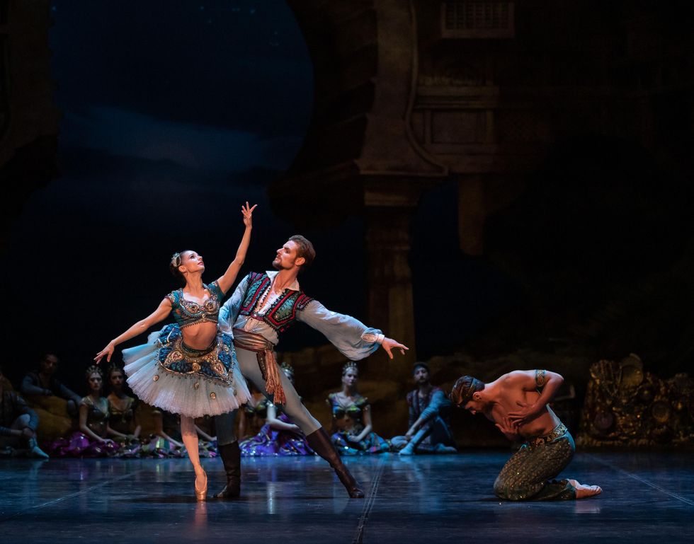 Wearing an ornate blue tutu and midriff-baring top, Maria Kochetkova balances in attitude derriere crois\u00e9 on her right leg whicle her parnter, who wears a pirate costume, stands in tendu and holds onto her waist. To their right, a shirtless dancer in blue sparkly pants kneels on the ground, bowing subserviently with his hands drawn to his chest.