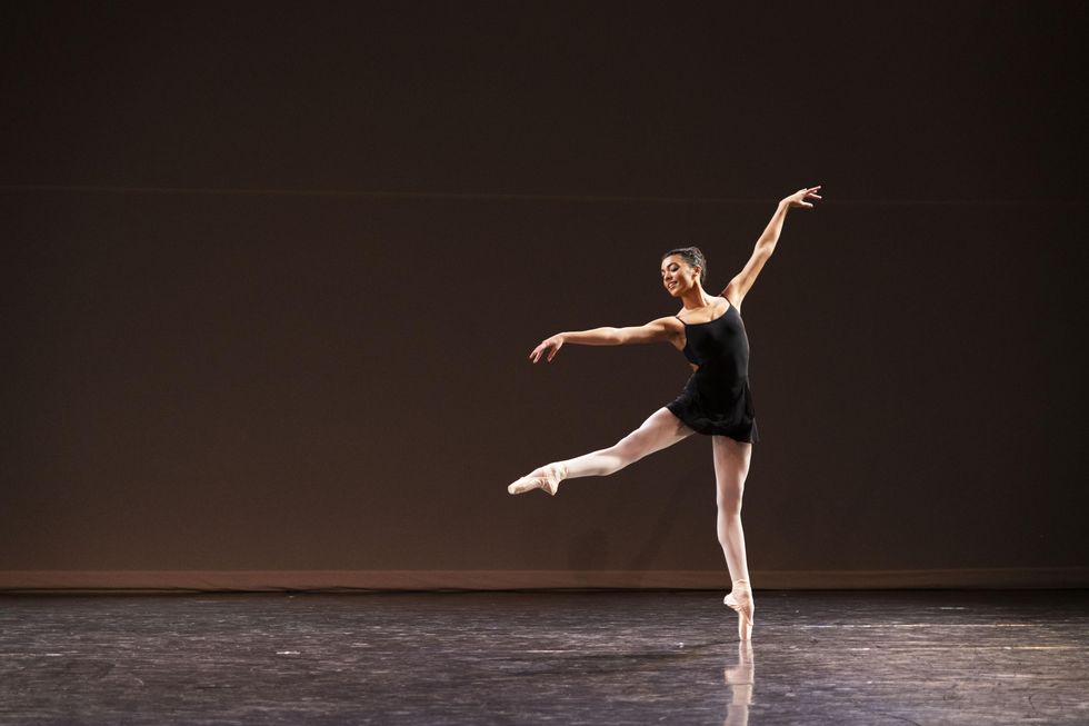Kyra Coco, a light skinned-Black ballet dancer, performs a sissone in second onto her left leg on pointe. Wearing a black spaghetti strap leotard and black skirt, she looks out over her right hand, which is stretched out to the side.