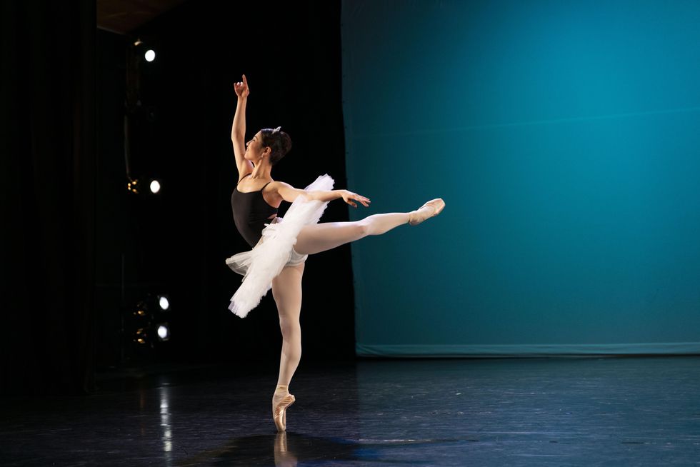Kotomi Yamada, a young Asian ballet dancer, performs a piquu00e9 attitude on pointe on her right leg and looks up toward her raised right arm. She wears a black camisole leotard and white practice tutu and dances on a black box stage in front of a blue backdrop.