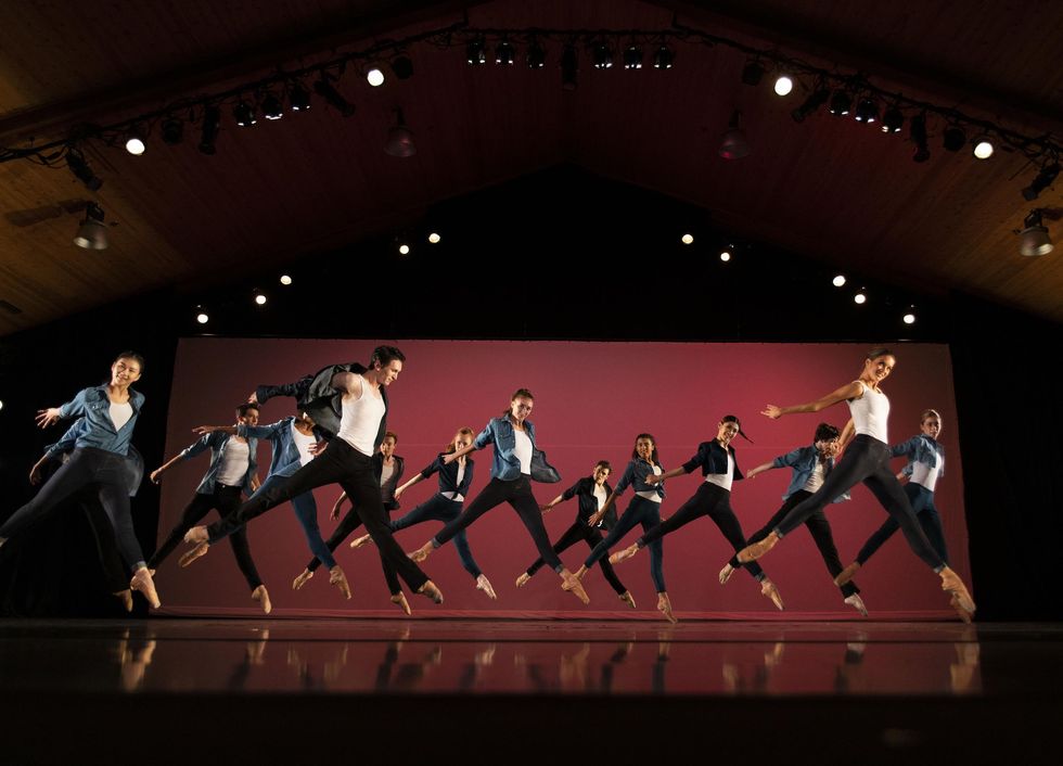 A large group of male and female dancers in black leggings, white T-shits and jean jackets do a sissone with their left legs in front and their arms thrown back. They jump in front of a red backdrop, cocking their heads towards the audience with big smiles.