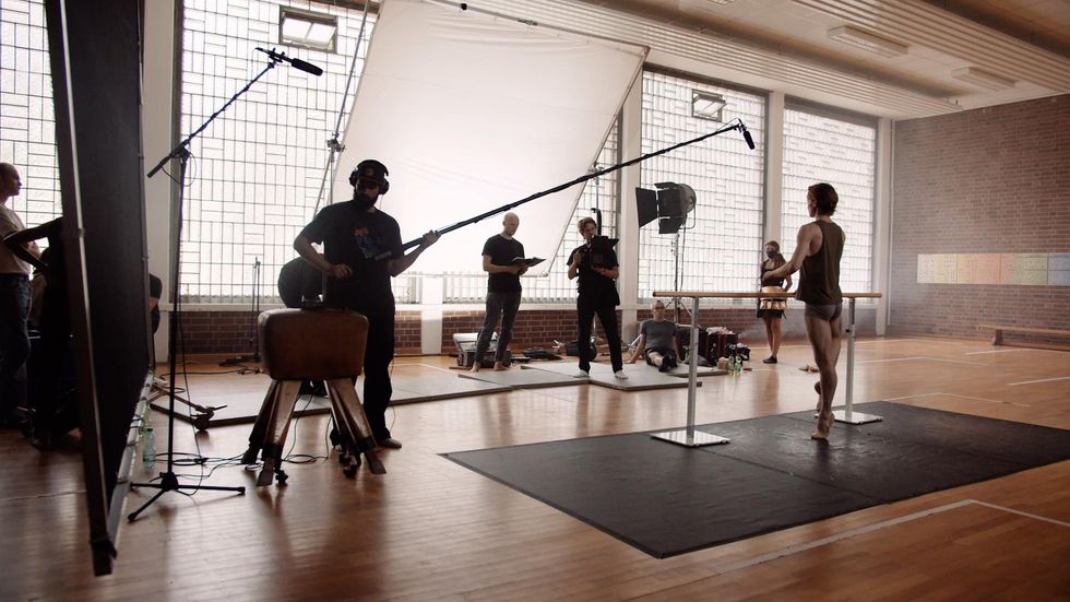 In a gymnasium set up with large boom lights and video cameras, Daniil Simkin balances in coup\u00e9 a the barre with his back away from the camera. He wears gray booty shorts, a green tank top and tan ballet slippers. Three crewmen and a woman in a patterned skirt stand in front of him.