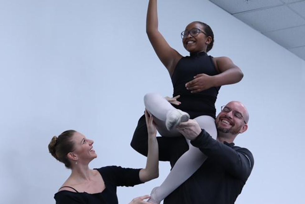 A young female ballet student in a black turtleneck leotard, pink tights and slippers and wearing glasses, sits on Francis Vayette's shoulder with her right leg in attitude and her left leg tucked under. Lauren Vayette touches her knee to make sure she's on balance and smiles up at her.