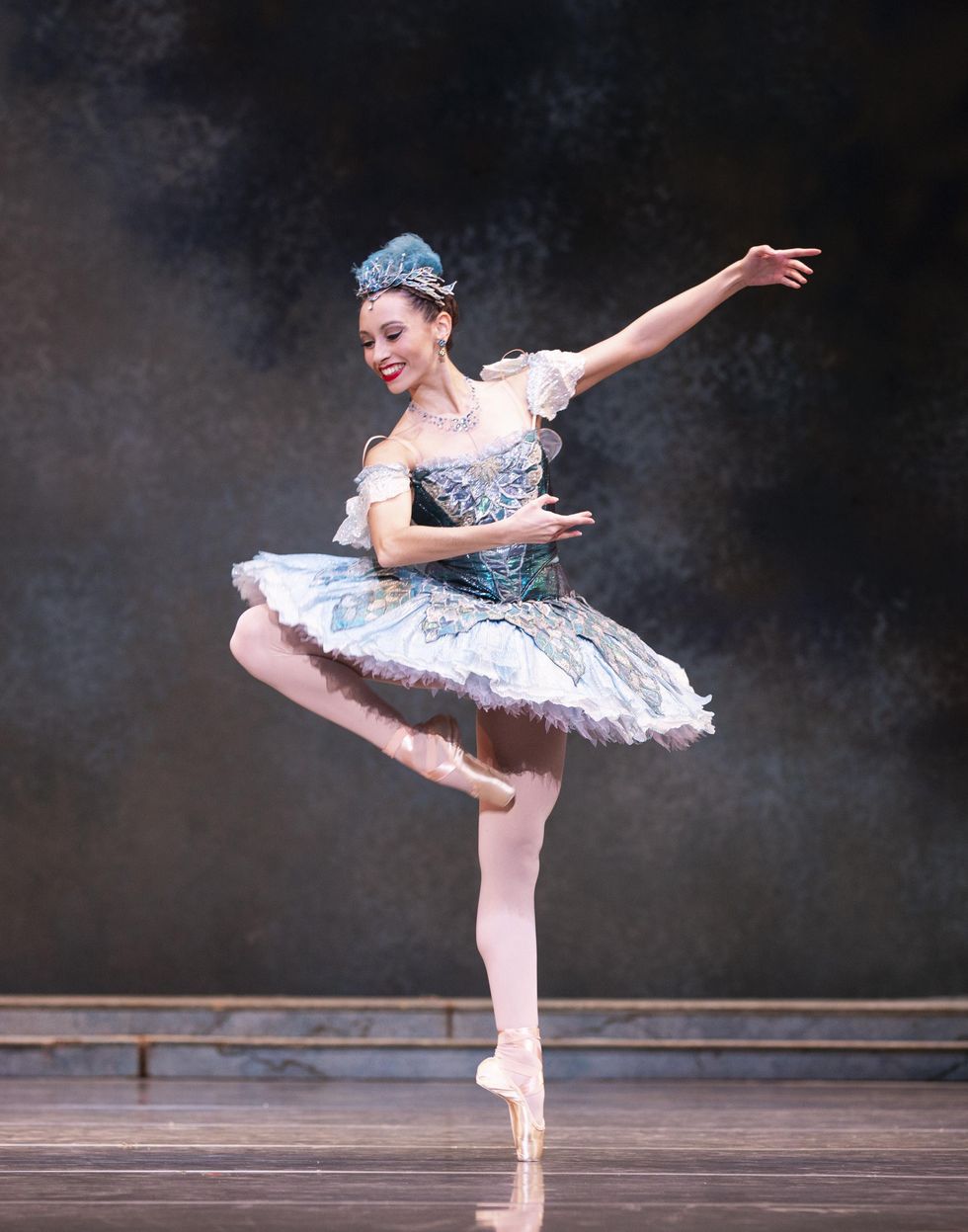 Sarah-Gabrielle Ryan does a piqu\u00e9 retir\u00e9 on her left foot and holds her arms in fourth position en evant, leaning her upper body over to look towards her right leg. She wears an ornate blue tutu with off-the-shoulder sleeves, a blue feathered headress and pink tights and pointe shoes.