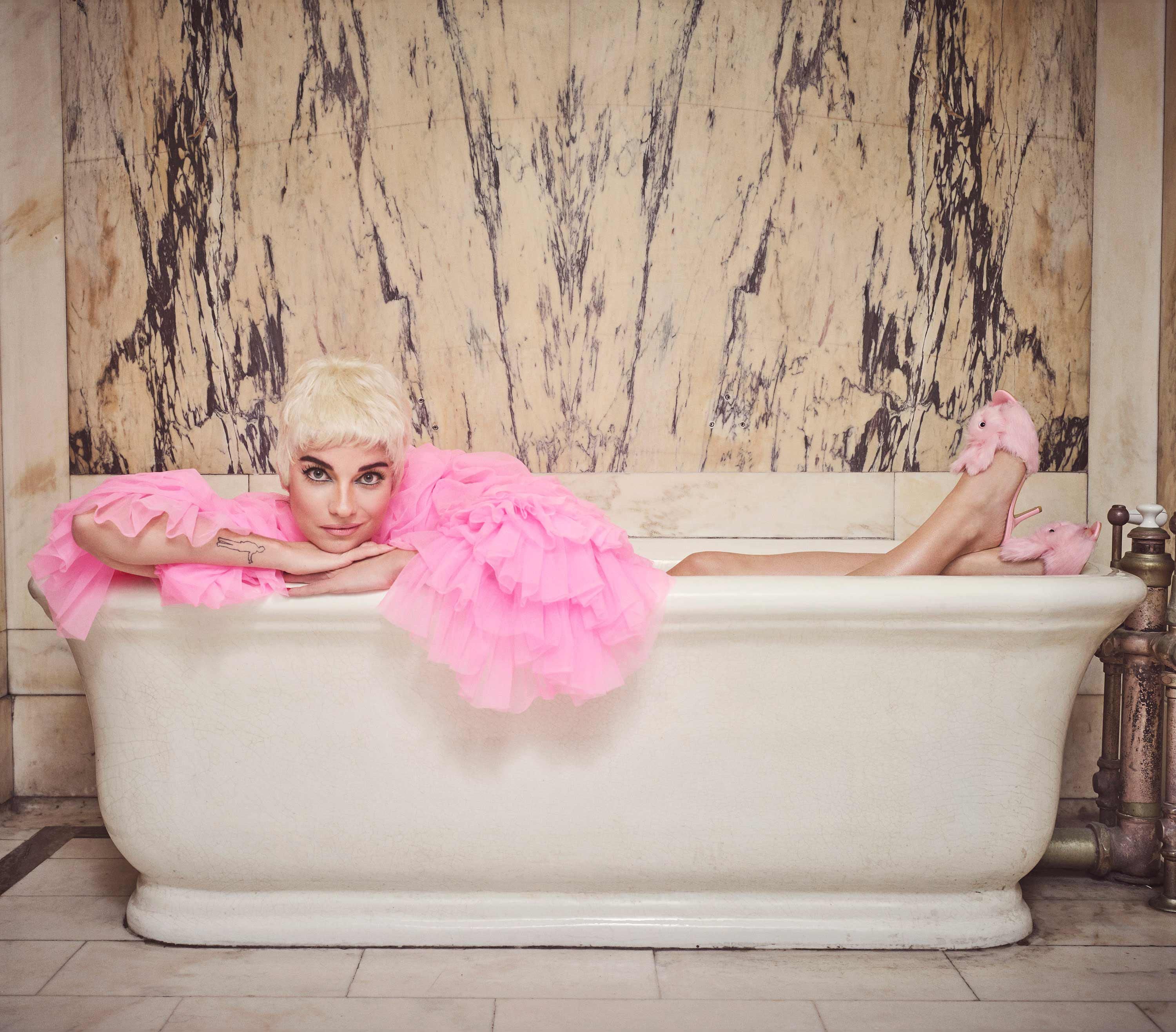 Annie Murphy poses in a tub wearing pink bunny slipper heels and a ruffled pink jacket