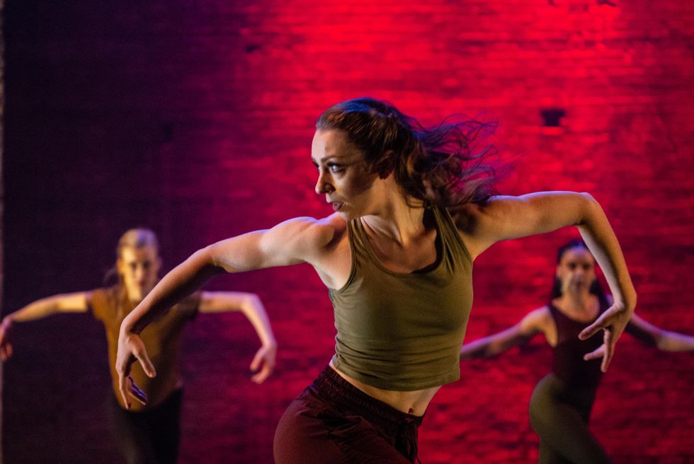 Zo\u00e9 Emilie Henrot looks sharply to the right and holds her arms out to the side with her elbows bent while twisting her lower body left at the hips. She wears an olive green tank top and burgundy tights, and is flanked by two female dancers behind her on either side doing the same pose. A red light glows on the brick wall behind them.