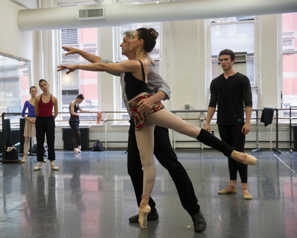 Luciana Paris, in a black leotard and short patterned skirt, stands in a low arabesque on pointe on her right leg and wraps her arm around Kevin McKenzie's left shoulder. McKenzie, in a blue shirt and black pants, holds her waist and does a small lunge to the right with his right arm extended out. Behind them in the studio, several male and female dancers look on.