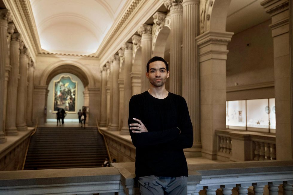 Wearing a black long-sleeved shirt and gray pants, Silas Farley crosses his arms and leans against a marble railing inside of a grand museum. He smiles slightly and seriously towards the camera.