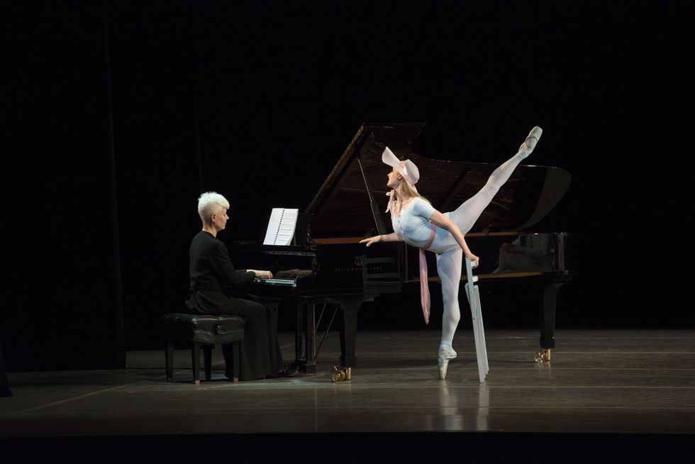 Ashley Ellis, dressed in a floppy hat and a blue leotard, blue tights and pointe shoes, presses down on a black grand piano onstage with her right hand and lifts her left leg high in arabesque allong\u00e9 while holding a folding chair in her left hand. She looks attentively toward the pianist, a woman with short white hair dressed all in black..
