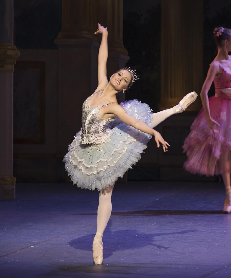 Wearing a sparkly pancake tutu and tiara, Ashley Ellis does a piqu\u00e9 attitude derriere crois\u00e9 on her left leg, lifting her right arm up and her left arm to the side. She arches her upper body out towards the audience and smiles brightly,.