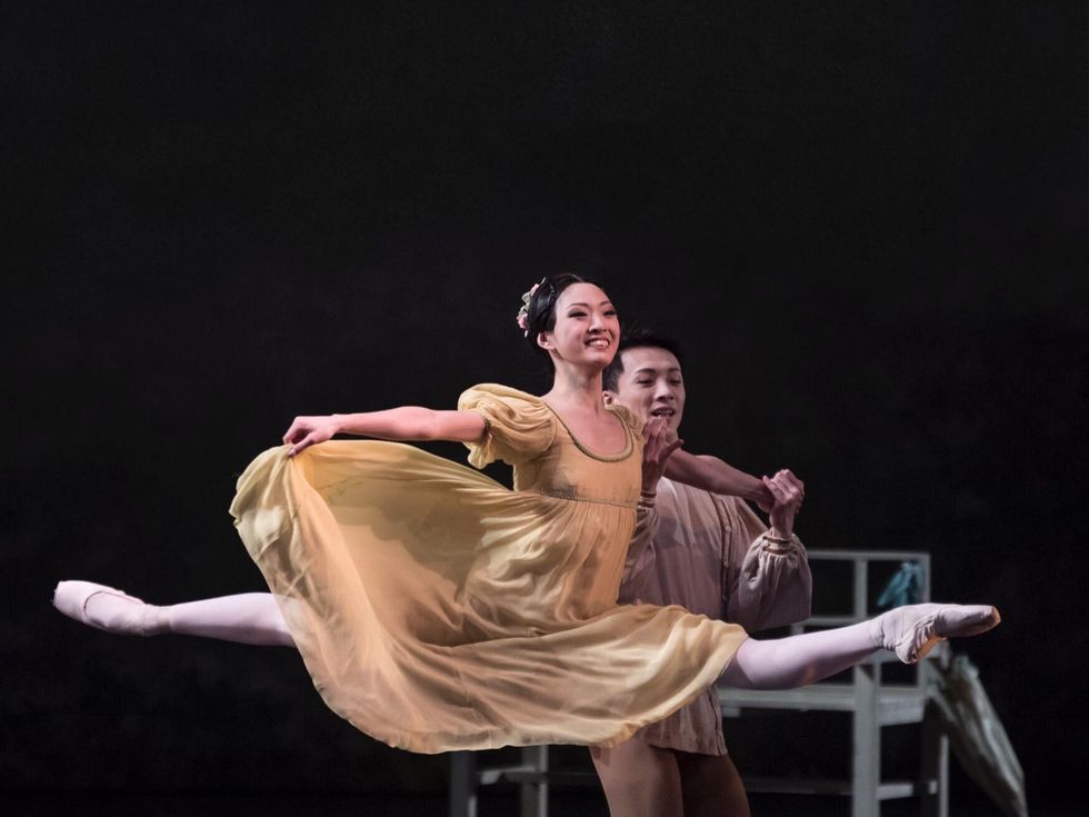 Patricia Zhou, wearing a big smile, a beige empire-waist dress and a floral headpiece, does a partnered grand jet\u00e9 to the left, holding onto her male partner's left hand with her own left hand. She holds her skirt out to the side with her right hand.