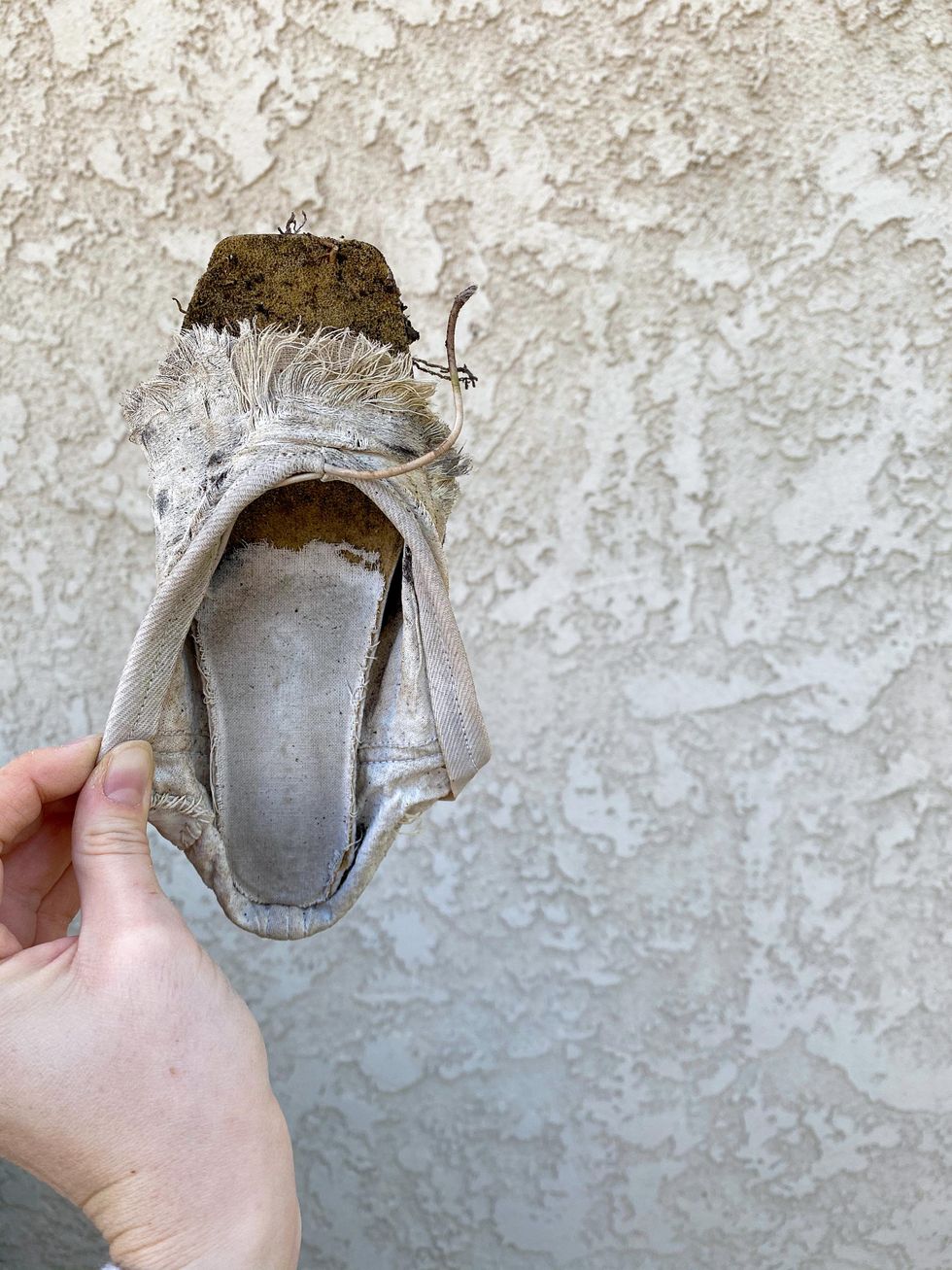 A hand holds up a disintegrated pointe show, with dirty, frayed satin, against a white cement wall.