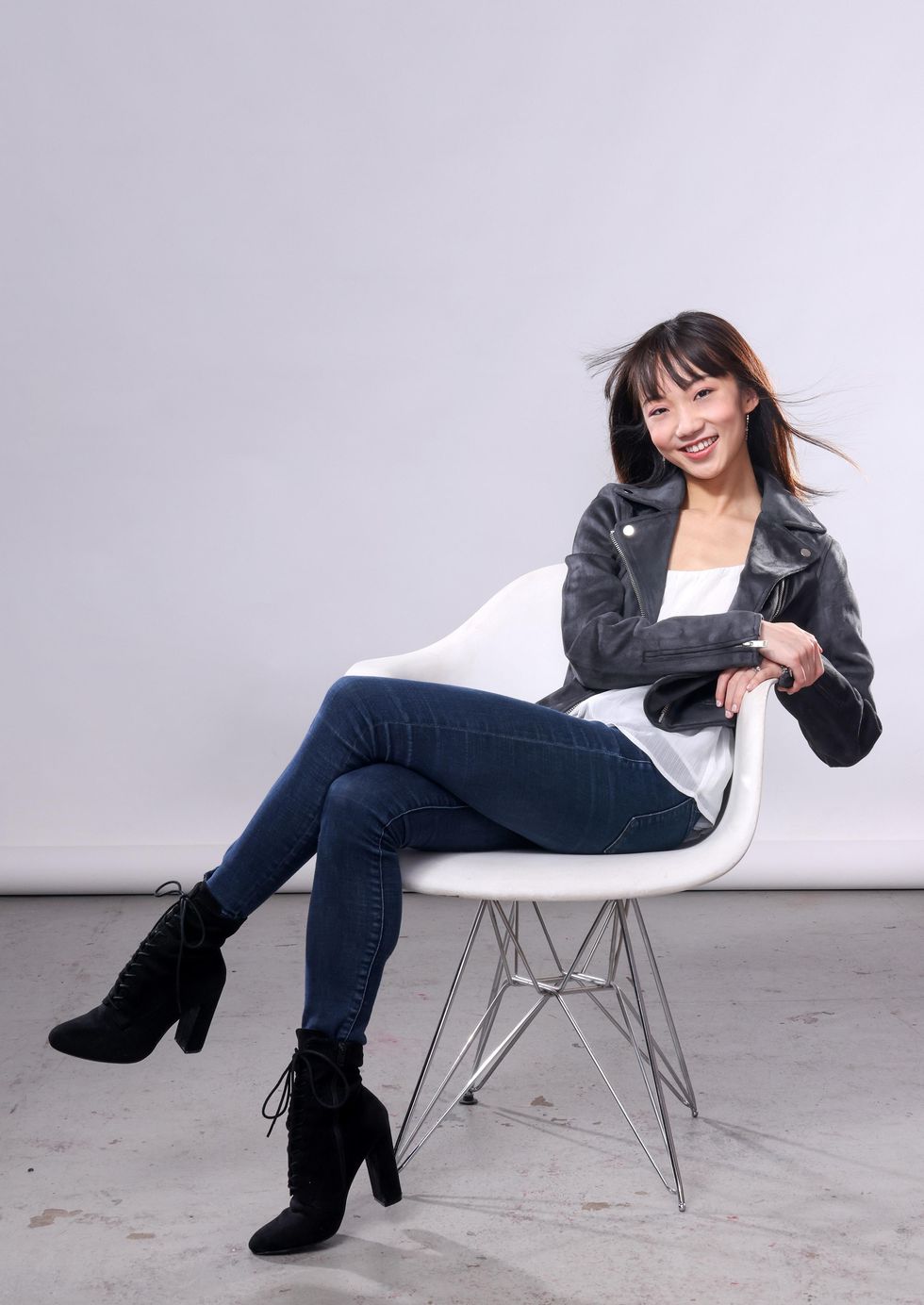 A smiling, confident Chisako Oga sits cross-legged in a white chair, her arms leaning on the chair's left arm. She wears stretch jeans, high-heeled black boots, a filmy white shirt and black leather jacket.